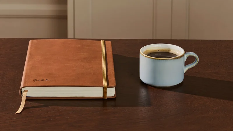 Lædernotesbog med bogmærkebånd ved siden af en kop kaffe